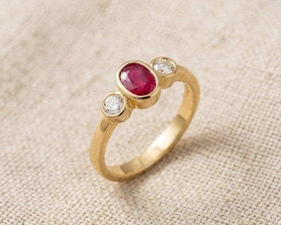 18ct yellow gold trilogy ring with a central oval-cut ruby and two side diamonds, set in bezels, on a light linen fabric background. Close-up studio photograph of an 18ct yellow gold ring featuring an oval ruby and two brilliant-cut diamonds. (Trilogy bezel setting).