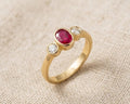18ct yellow gold trilogy ring with a central oval-cut ruby and two side diamonds, set in bezels, on a light linen fabric background. Close-up studio photograph of an 18ct yellow gold ring featuring an oval ruby and two brilliant-cut diamonds. (Trilogy bezel setting).