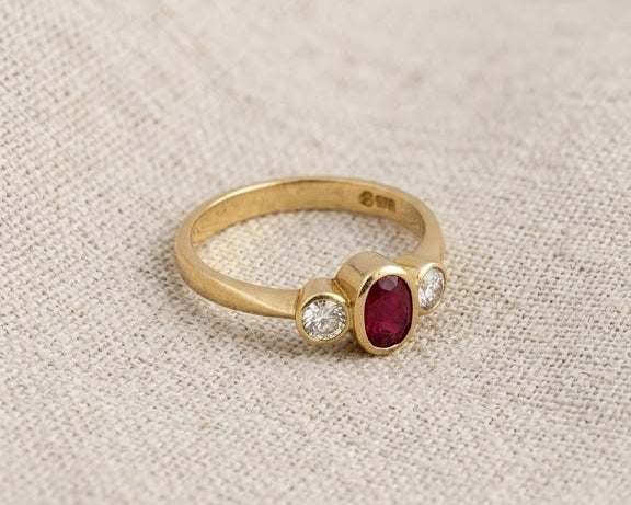 18ct yellow gold trilogy ring with a central oval-cut ruby and two side diamonds, set in bezels, on a light linen fabric background. Close-up studio photograph of an 18ct yellow gold ring featuring an oval ruby and two brilliant-cut diamonds. (Trilogy bezel setting).