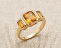 A detailed high-resolution product photo of a yellow gold three-stone ring with bezel-set emerald-cut citrines, showcasing clear, deep golden stones against a woven light fabric.