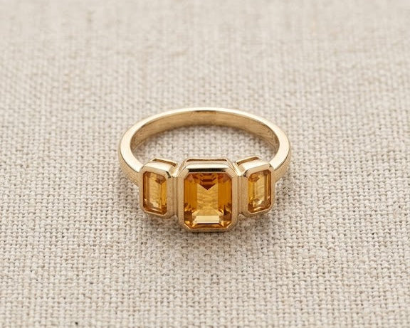 A detailed high-resolution product photo of a yellow gold three-stone ring with bezel-set emerald-cut citrines, showcasing clear, deep golden stones against a woven light fabric.