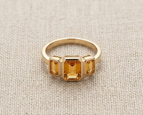 A detailed high-resolution product photo of a yellow gold three-stone ring with bezel-set emerald-cut citrines, showcasing clear, deep golden stones against a woven light fabric.