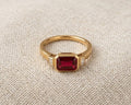 Modern emerald-cut deep red garnet solitaire ring with two flanking baguette-cut diamond accents, set in a high-polished yellow gold bezel setting, resting on a textured natural flax-colored linen fabric with soft, flattering studio lighting. The profile view showcases the substantial depth, rich velvety color, and crisp step facets of the genuine rectangular garnet gemstone.