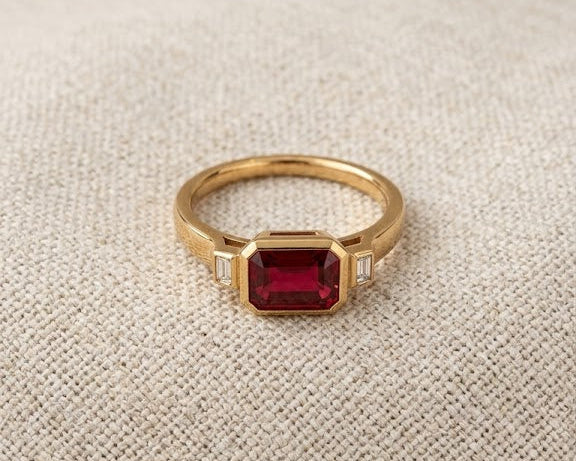 Modern emerald-cut deep red garnet solitaire ring with two flanking baguette-cut diamond accents, set in a high-polished yellow gold bezel setting, resting on a textured natural flax-colored linen fabric with soft, flattering studio lighting. The profile view showcases the substantial depth, rich velvety color, and crisp step facets of the genuine rectangular garnet gemstone.