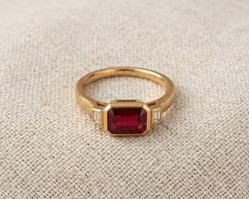 Modern emerald-cut deep red garnet solitaire ring with two flanking baguette-cut diamond accents, set in a high-polished yellow gold bezel setting, resting on a textured natural flax-colored linen fabric with soft, flattering studio lighting. The profile view showcases the substantial depth, rich velvety color, and crisp step facets of the genuine rectangular garnet gemstone.
