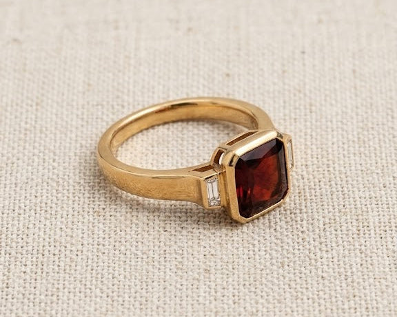 Modern emerald-cut deep red garnet solitaire ring with two flanking baguette-cut diamond accents, set in a high-polished yellow gold bezel setting, resting on a textured natural flax-colored linen fabric with soft, flattering studio lighting. The profile view showcases the substantial depth, rich velvety color, and crisp step facets of the genuine rectangular garnet gemstone.
