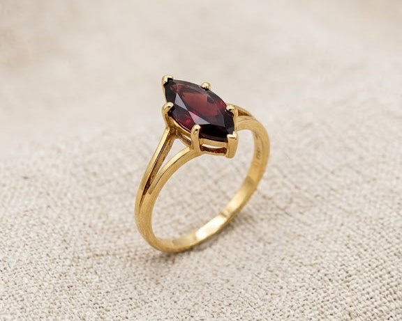 An exquisite 9ct yellow gold ring featuring a deep crimson marquise-cut garnet in a secure prong setting, resting on a textured light beige linen fabric surface under professional studio lighting.