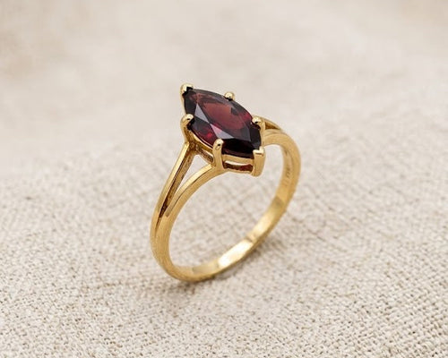 An exquisite 9ct yellow gold ring featuring a deep crimson marquise-cut garnet in a secure prong setting, resting on a textured light beige linen fabric surface under professional studio lighting.