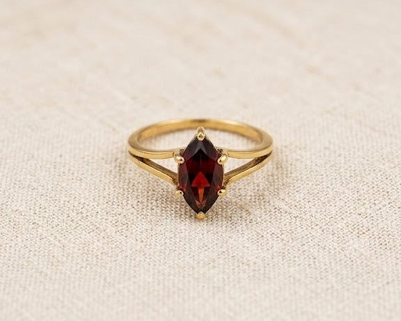 An exquisite 9ct yellow gold ring featuring a deep crimson marquise-cut garnet in a secure prong setting, resting on a textured light beige linen fabric surface under professional studio lighting.