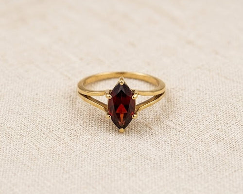 An exquisite 9ct yellow gold ring featuring a deep crimson marquise-cut garnet in a secure prong setting, resting on a textured light beige linen fabric surface under professional studio lighting.