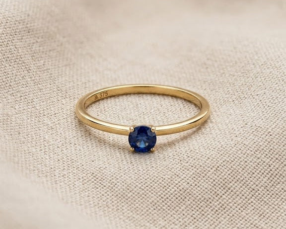 A minimalist 9ct yellow gold sapphire solitaire ring with a round deep blue sapphire, claw-set on a dainty gold band, displayed on a textured light linen fabric with professional studio lighting.
