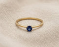 A minimalist 9ct yellow gold sapphire solitaire ring with a round deep blue sapphire, claw-set on a dainty gold band, displayed on a textured light linen fabric with professional studio lighting.