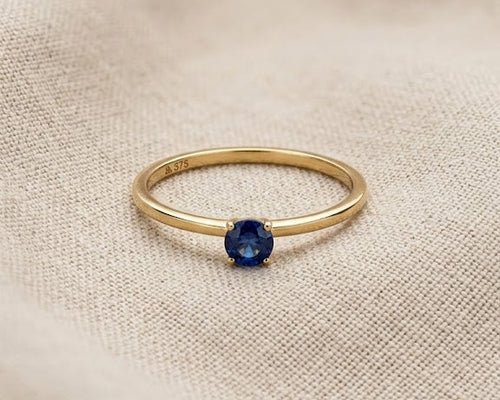 A minimalist 9ct yellow gold sapphire solitaire ring with a round deep blue sapphire, claw-set on a dainty gold band, displayed on a textured light linen fabric with professional studio lighting.