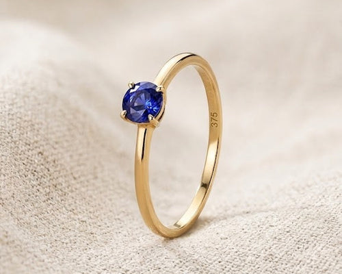 A minimalist 9ct yellow gold sapphire solitaire ring with a round deep blue sapphire, claw-set on a dainty gold band, displayed on a textured light linen fabric with professional studio lighting.