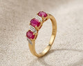 A detailed studio photograph of a 9ct yellow gold ring with three oval-cut red rubies and four small white diamonds on a light beige linen fabric surface. This jewelry piece features a split-shank design with the diamonds accenting the rubies. Ideal for a woman's engagement or fashion accessory, the ring is presented in a well-lit, professional setting.