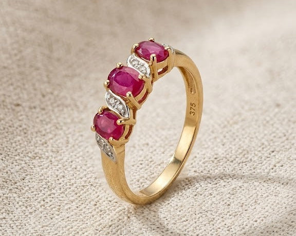 A detailed studio photograph of a 9ct yellow gold ring with three oval-cut red rubies and four small white diamonds on a light beige linen fabric surface. This jewelry piece features a split-shank design with the diamonds accenting the rubies. Ideal for a woman's engagement or fashion accessory, the ring is presented in a well-lit, professional setting.