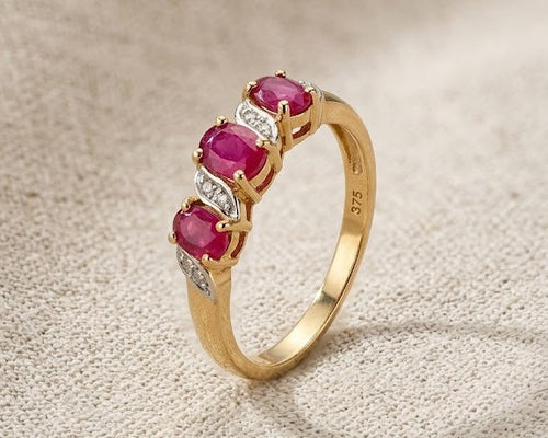 A detailed studio photograph of a 9ct yellow gold ring with three oval-cut red rubies and four small white diamonds on a light beige linen fabric surface. This jewelry piece features a split-shank design with the diamonds accenting the rubies. Ideal for a woman's engagement or fashion accessory, the ring is presented in a well-lit, professional setting.
