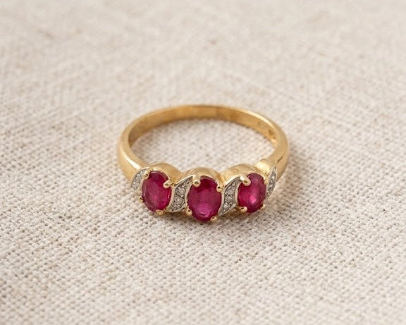 A detailed studio photograph of a 9ct yellow gold ring with three oval-cut red rubies and four small white diamonds on a light beige linen fabric surface. This jewelry piece features a split-shank design with the diamonds accenting the rubies. Ideal for a woman's engagement or fashion accessory, the ring is presented in a well-lit, professional setting.