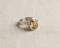 Oval-cut yellow citrine solitaire ring in a sterling silver bezel setting, resting on a textured linen fabric background. Angled.