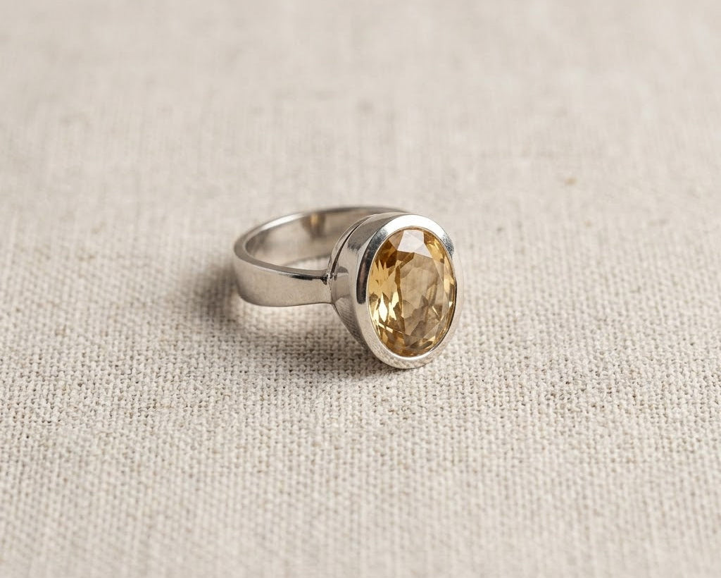 Oval-cut yellow citrine solitaire ring in a sterling silver bezel setting, resting on a textured linen fabric background. Angled.