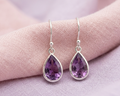 Pair of pear-cut purple amethyst drop earrings in sterling silver bezel settings, draped over textured pink and purple linen fabric.