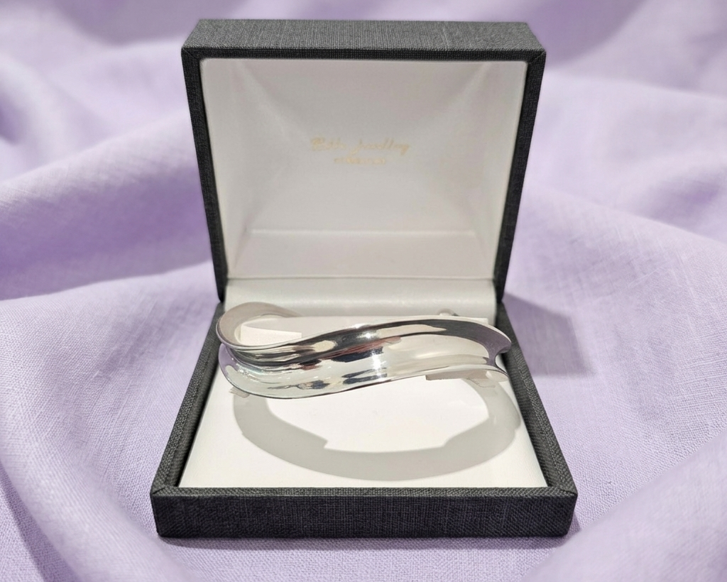 Sculptural sterling silver wide-wave bangle with a high-polished mirror finish, displayed in a black jewelry box on a textured lavender linen background.
