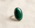 Large oval cabochon-cut green malachite gemstone with natural banding patterns, set in a polished sterling silver bezel ring, displayed on a light linen fabric background. Angled view.