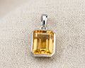 Modern emerald-cut citrine solitaire pendant set in a high-polished sterling silver bezel setting, resting on a textured light linen fabric with soft studio lighting. The pendant shows the deep golden-yellow clarity of the genuine citrine gemstone and its distinct step facets.
