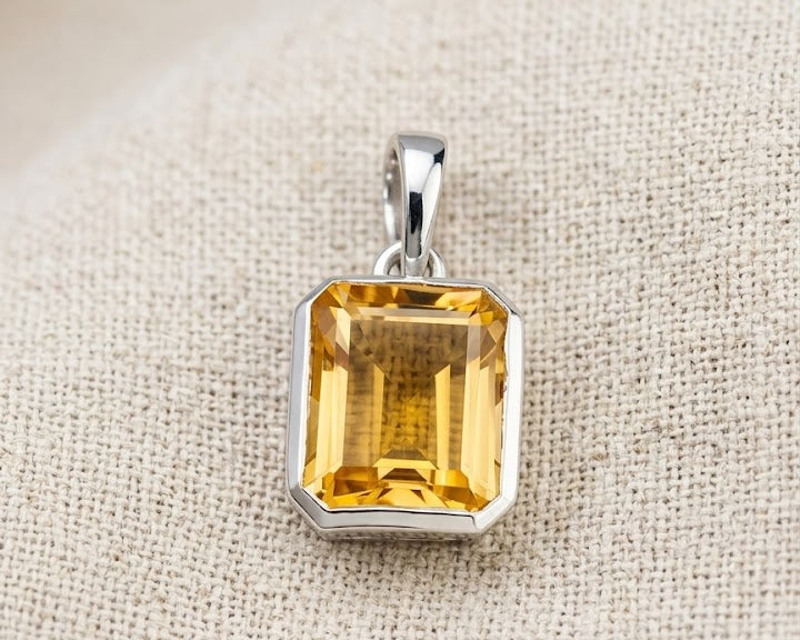 Modern emerald-cut citrine solitaire pendant set in a high-polished sterling silver bezel setting, resting on a textured light linen fabric with soft studio lighting. The pendant shows the deep golden-yellow clarity of the genuine citrine gemstone and its distinct step facets.
