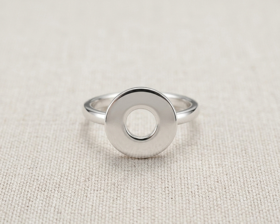 A highly polished sterling silver geometric ring with a prominent, flat circular face and a precise central hole, resting centered on a textured, light-oatmeal linen background.