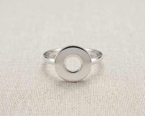 A highly polished sterling silver geometric ring with a prominent, flat circular face and a precise central hole, resting centered on a textured, light-oatmeal linen background.