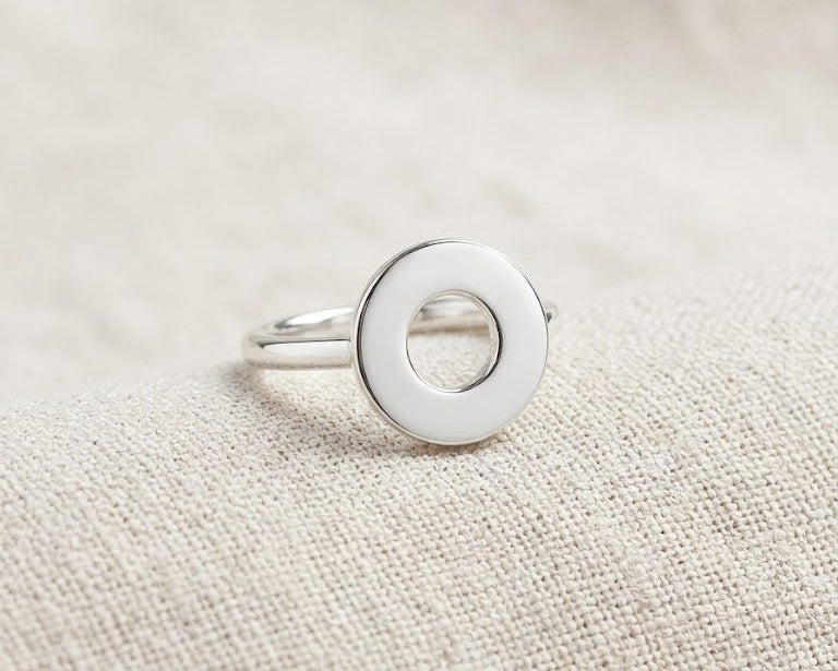 A highly polished sterling silver geometric ring with a prominent, flat circular face and a precise central hole, resting centered on a textured, light-oatmeal linen background with soft studio lighting. Angled view.