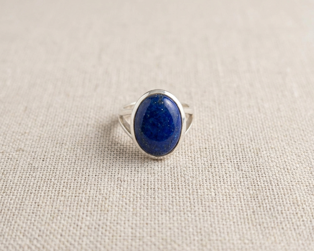 Large oval cabochon-cut deep blue Lapis Lazuli ring with natural golden pyrite inclusions, set in a polished sterling silver bezel band on a light linen background.  