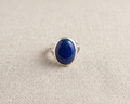 Large oval cabochon-cut deep blue Lapis Lazuli ring with natural golden pyrite inclusions, set in a polished sterling silver bezel band on a light linen background.  