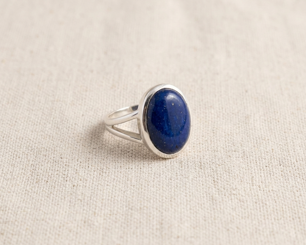 Large oval cabochon-cut deep blue Lapis Lazuli ring with natural golden pyrite inclusions, set in a polished sterling silver bezel band on a light linen background.  Angled view.