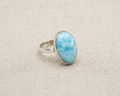 Oval cabochon Larimar gemstone ring with light blue and white sea-foam patterns, set in an adjustable sterling silver band, displayed on a neutral background. Angled view.