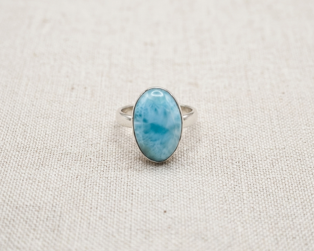 Oval cabochon Larimar gemstone ring with light blue and white sea-foam patterns, set in an adjustable sterling silver band, displayed on a neutral background.