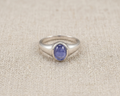 Oval-cut purple-blue tanzanite gemstone set in a polished sterling silver signet-style ring, displayed on a neutral linen fabric background with soft studio lighting.