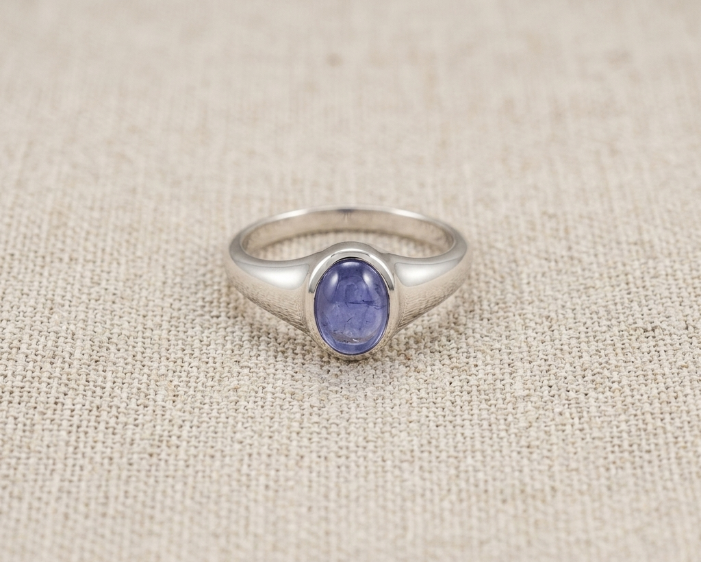 Oval-cut purple-blue tanzanite gemstone set in a polished sterling silver signet-style ring, displayed on a neutral linen fabric background with soft studio lighting.