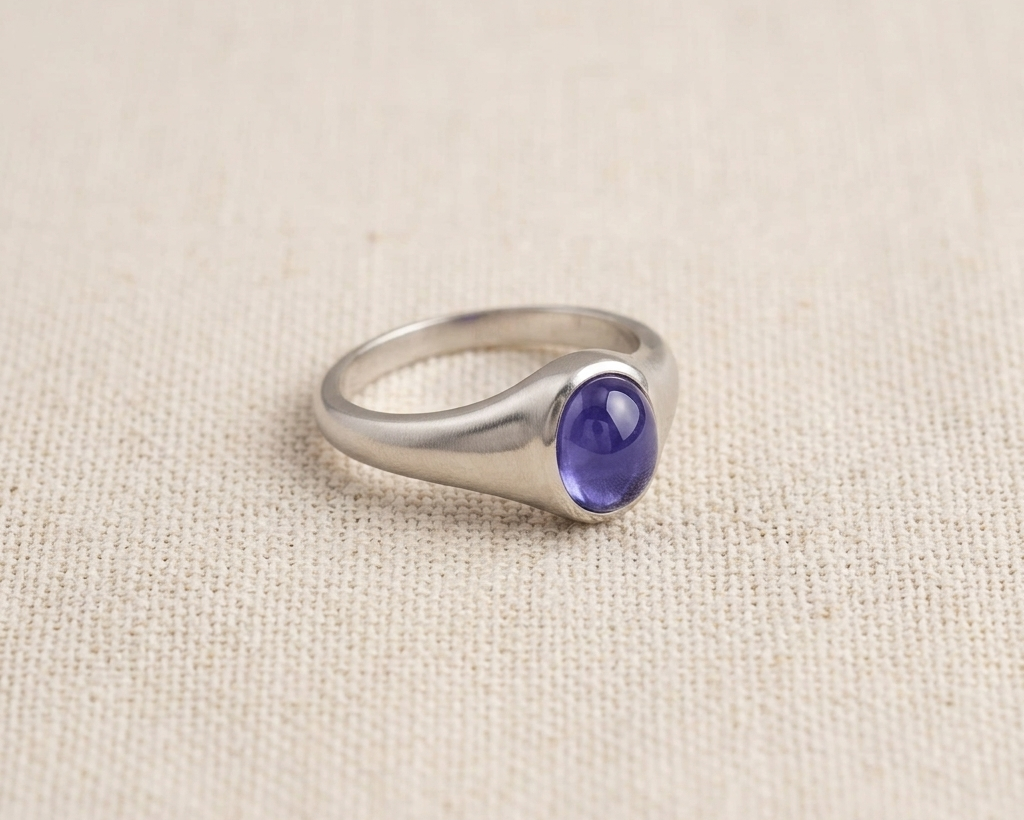 Oval-cut purple-blue tanzanite gemstone set in a polished sterling silver signet-style ring, displayed on a neutral linen fabric background with soft studio lighting. Angled.
