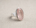 Oval faceted rose-cut pink rose quartz gemstone set horizontally in a polished sterling silver bezel band ring, resting on a textured light beige linen fabric with soft studio lighting. Angled view
