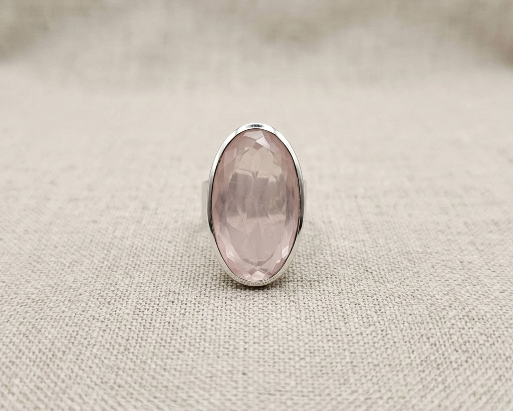 Oval faceted rose-cut pink rose quartz gemstone set horizontally in a polished sterling silver bezel band ring, resting on a textured light beige linen fabric with soft studio lighting.