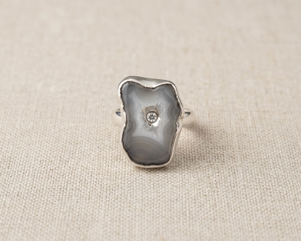 Irregular natural organic grey and white Seafoam Agate slice ring with a round bezel-set blue topaz center stone, all set in sterling silver, displayed on a light linen fabric.