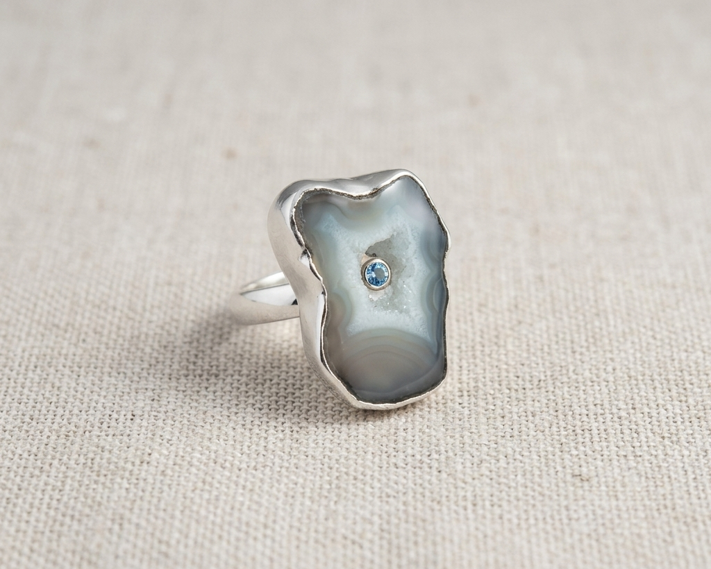 Irregular natural organic grey and white Seafoam Agate slice ring with a round bezel-set blue topaz center stone, all set in sterling silver, displayed on a light linen fabric. Angled view.