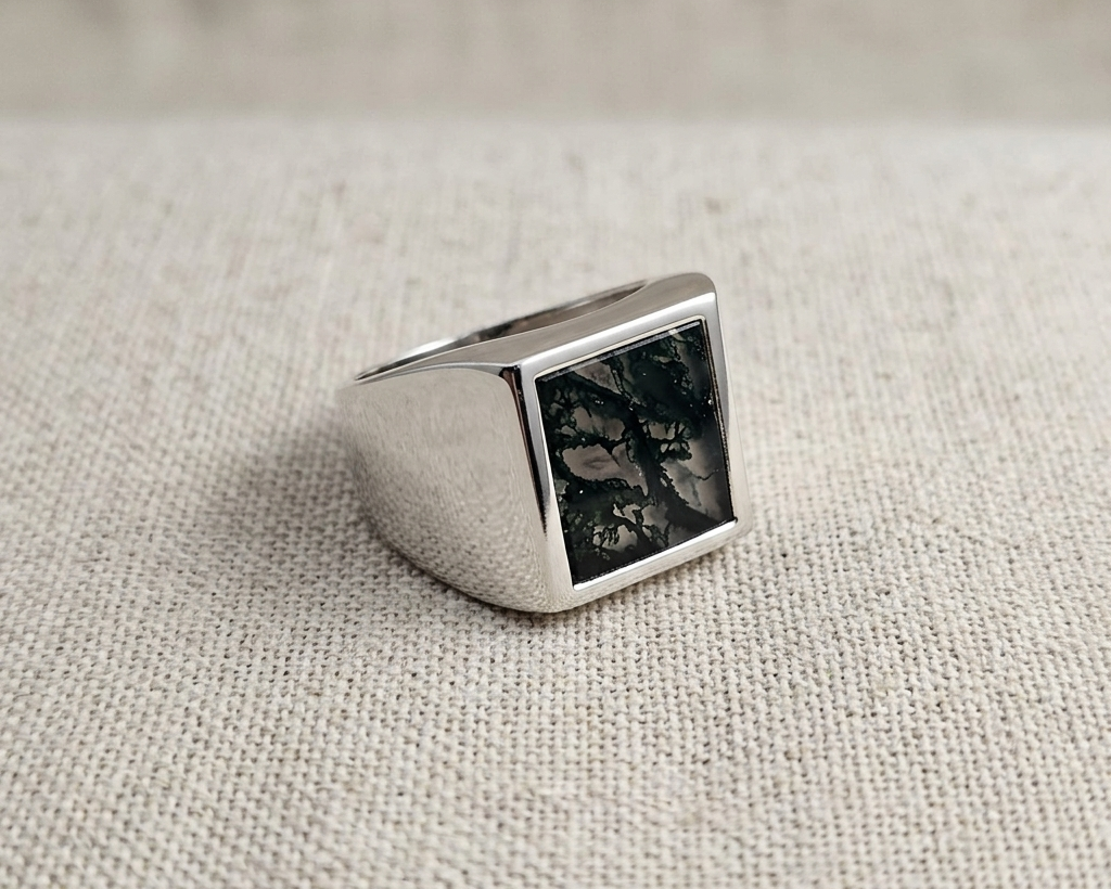 Polished sterling silver Scribe-style signet ring with a natural, unique moss agate stone panel featuring dark green dendritic inclusions, displayed on a light linen fabric.  Angled view.