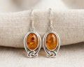 A matched pair of oval genuine Baltic Amber drop earrings set in detailed .925 sterling silver Celtic knotwork settings, featuring comfortable hooks for pierced ears.