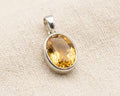 Faceted oval citrine gemstone pendant in a polished sterling silver bezel setting, displayed on a light linen fabric background.