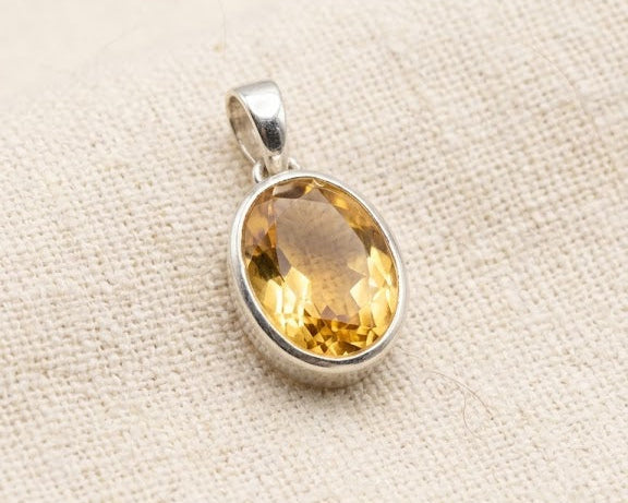 Faceted oval citrine gemstone pendant in a polished sterling silver bezel setting, displayed on a light linen fabric background.
