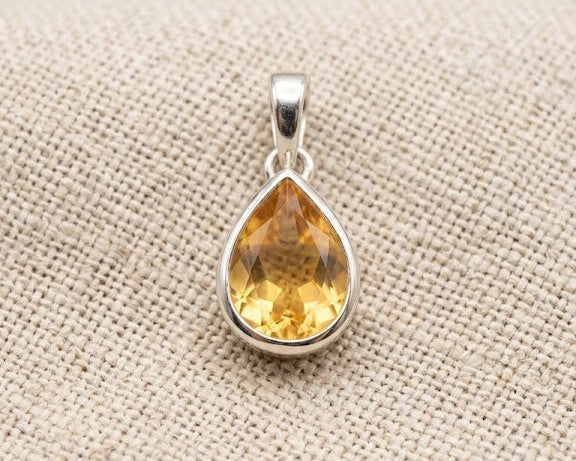 Professional photo of a polished sterling silver pendant featuring a vibrant pear-cut citrine gemstone, resting on a textured linen surface with soft studio lighting.