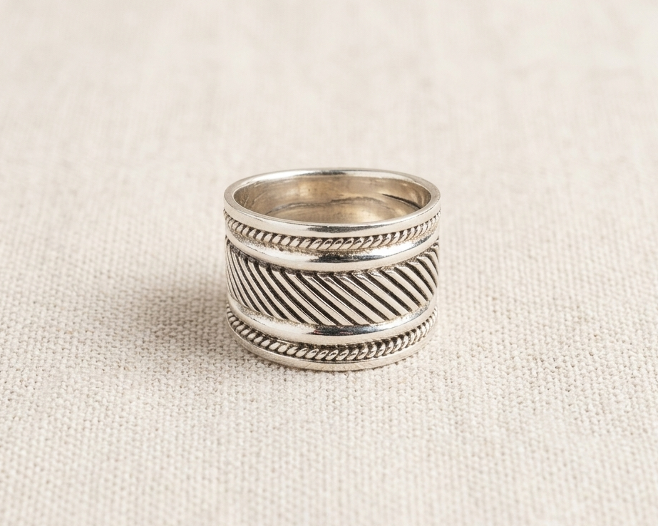 Substantial sterling silver wide band ring featuring an intricate oxidized diagonal woven pattern bordered by delicate rope-twist edges, displayed on a textured light linen background.
